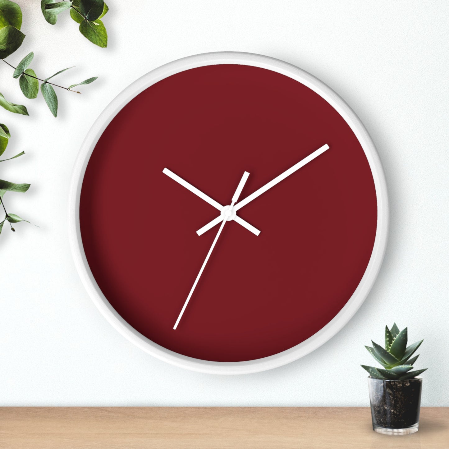 White circular wooden framed clock with dark red background and white hands with leaves on upper left corner and small cactus on bottom right against a white background.