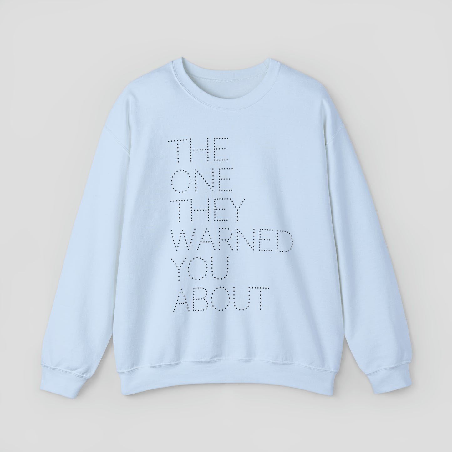Front view of light blue sweatshirt with with the words "The One They Warned You About" in capitalized dotted text in center, against a gray background.